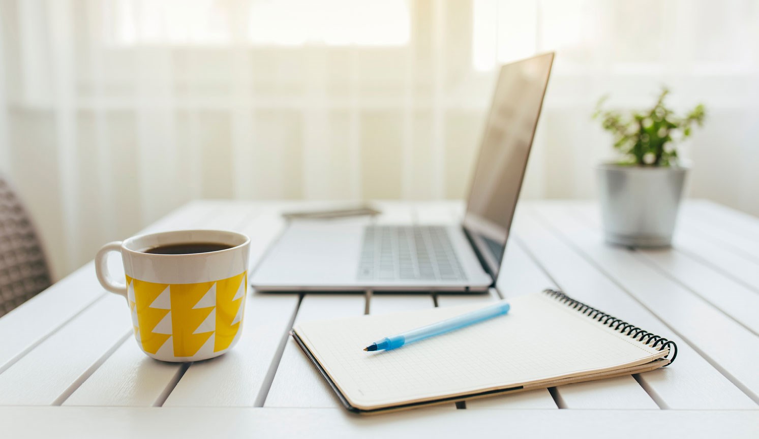 laptop, notepad, coffee in yellow mug on sunlit desk